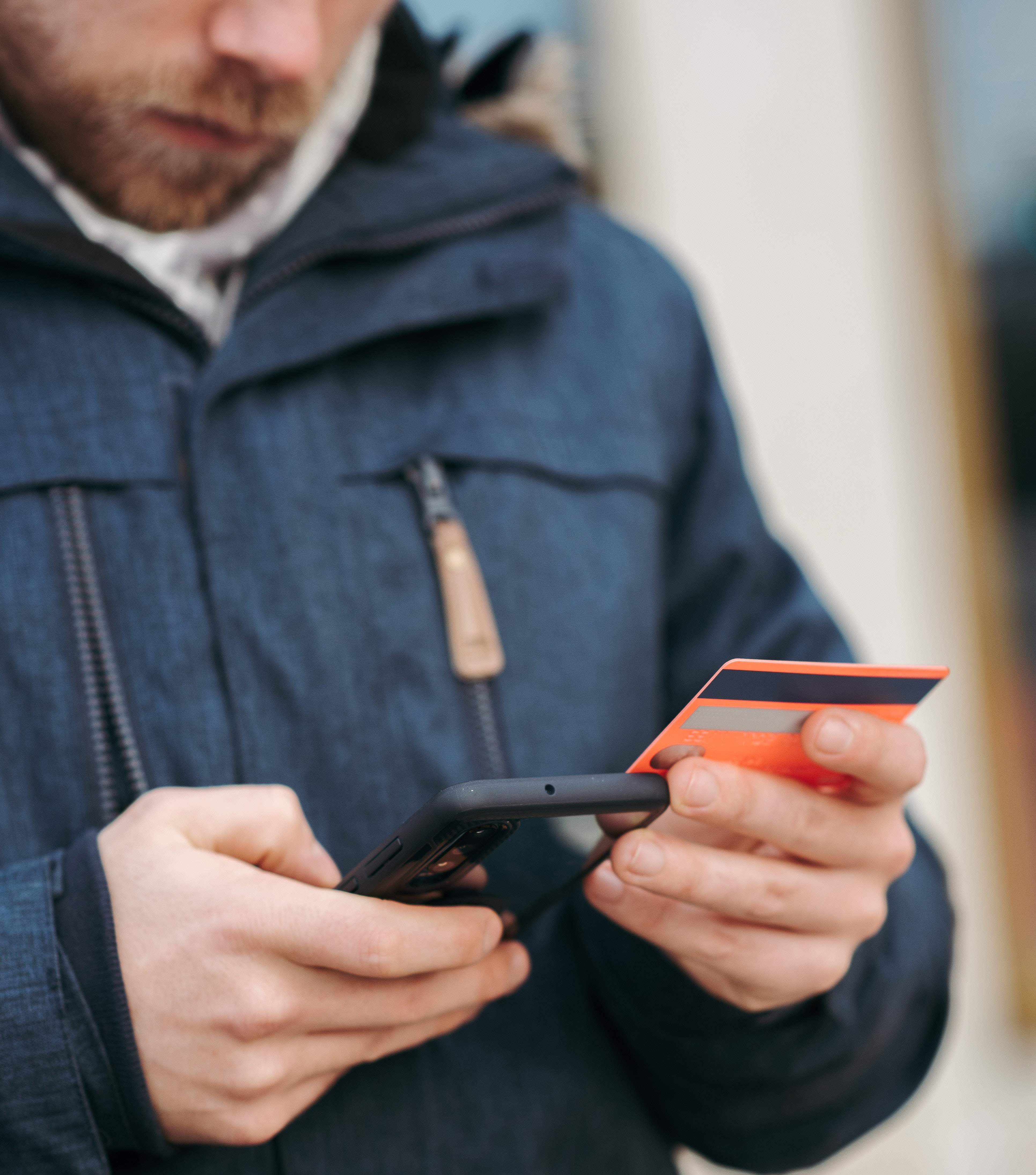 Foto von einer weißen Person in einer Winterjacke. In der rechten Hand ist ein Smartphone, in der linken eine Kreditkarte.